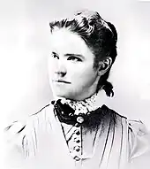 A washed-out monochrome photograph portrait of a young woman with shoulders squared to the camera, the head turned to the left, a neutral-colored dress topped by a dark ruff and white lace, the woman's dark hair pulled back behind her ears to fall down below her collar