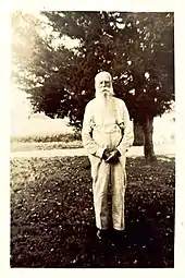 A black and white image (c.1900s) of an elderly man with white hair and a long beard dressed in a long-sleeved and suspenders standing in front of a tree.