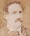 Faded sepia photograph showing the head and shoulders of a man with dark, wavy hair, mustache and wearing a dark coat, white shirt with wingtip collar and dark cravat