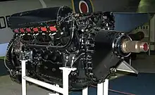 A front right view of a large, black-painted, piston aircraft engine with a prominent propeller shaft. A camouflaged military aircraft is parked behind.