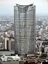 Aerial view of one curved side of a tall building set in front of a cityscape; the facade is symmetrical and divided into three vertical sections: both identical sections on either side are divided by a section in the center that turns into a large atrium window as it nears the building's roof