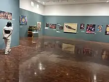 foto panorámica de una sala en el museo durante la exposición "otros muralismos", encuentras una esquina de la sala en la que se exponen al menos 15 obras poco detalladas de la sala entera, es una sala color verde con piso cafe madera y techo blanco, en las paredes están los cuadros que retratan los trabajos de los artistas