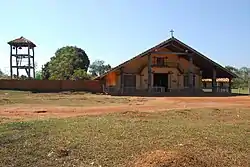 Un campanario de madera y una iglesia desde una vista frontal más allá de un área de césped. Una cruz se coloca en la parte superior del techo.