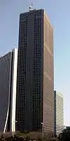 Ground-level view of a brown, rectangular high-rise; the window placement creates several horizontal bands on one side and one vertical stripe on the other