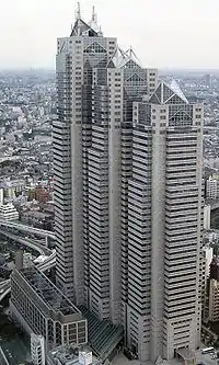 Aerial view of a building's two beige facades with horizontal rows of windows set in front of a cityscape; the building is composed of three adjoined towers of differing heights, each capped off with triangular glass structures