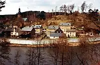 View of a small village with houses and a church, located in a wooded valley, with a river in the foreground. A high concrete wall separates the village from the river.