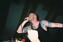 Vocalist Andrew Schwab performs at a concert. He has shaved his head and is wearing a black sleeveless shirt that displays his tattoos.