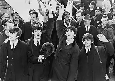 Fotografía en blanco y negro de los Beatles saludando frente a una multitud con un par de escaleras de avión en el fondo