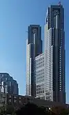 Ground-level view of a grey, window-dotted front building facade set in front of a dark blue sky; as the building rises, two towers break off on both sides