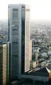 Aerial view of a building's two white, window-dotted facades set in front of a cityscape; its corners are beveled and are made completely of glass. A brown, horizontal-window-stripped building rising to half of the building's height is located immediately to its left