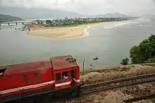 In the foreground, a red diesel locomotive is travelling up a mountain against the backdrop of Lang Co Beach and the sea. In the background are the mountains.