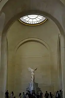 Escultura en mármol de mujer alada de pie vestida con toga, no posee brazos ni cabeza.