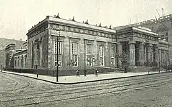 Un edificio de estilo neoegipcio en la esquina de una calle; hay columnas dóricas en la fachada a la derecha de la imagen. Hay hombres en la calle, y se está trabajando con grúas, en un edificio adyacente fuera de cuadro a la derecha.