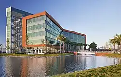 Edificio corporativo moderno de cinco niveles con fachada vidriada, rodeado por palmeras, frente a una laguna artificial.