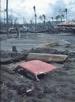 Rabaul luego de la erupción volcánica del 1994. En la fotografía se aprecia un auto rojo tapado casi en su totalidad, de lo que parece ser ceniza y barro, apenas viéndose el techo, al fondo se ven casas destruidas y palmeras de las que solo quedo el tronco.