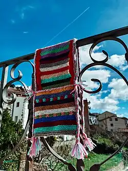 Alforja de lana y fieltro con franjas de muchos colores y pompones en los costados. Se encuentra sobre la barandilla del balcón de una casa