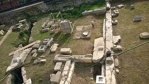 El frente de la Estoa Basileos en toda su longitud (a la derecha de la imagen), vista desde su costado norte. En primer plano, el ala norte (cuerpo lateral saliente). El «Camino del oeste» y el «Desagüe grande», parcialmente cubierto por losas (a la izquierda de la imagen), dispuestos en el frente del edificio.