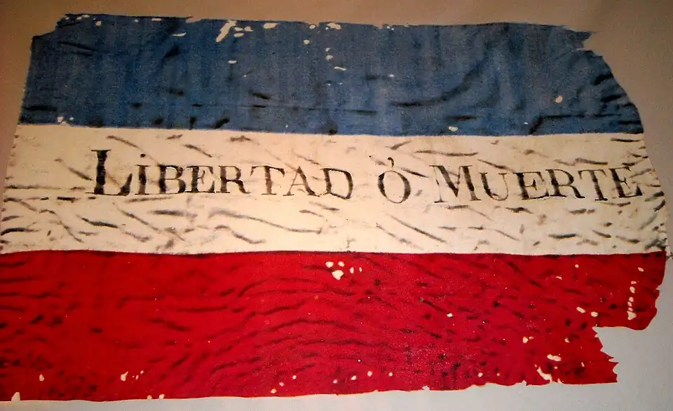 Reproducción fotográfica de la bandera original de los “Treinta y Tres Orientales” “Siendo una consecuencia necesaria del rango de la independencia y libertad que recobrado de hecho y de derecho la Provincia Oriental, fijar el pabellón que debe señalar su Ejercito y flamear en los pueblos de su territorio, se declara por tal, el que tiene por admitido, compuesto de las tres franjas horizontales celeste, blanca y punzo” Villa de San Fernando de la Florida 25 de agosto de 1825. Ubicación de este material, Museo del Cerro de Montevideo-Uruguay. Abril. 2014