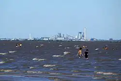 Fábrica a lo lejos, del otro lado del río. Dos bañistas caminan por el río.