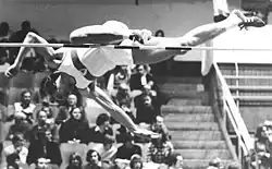 Un hombre con uniforme de atleta se lanza hacia delante, boca abajo sobre una barra de salto de altura. Su pierna derecha está estirada, paralela a la barra, mientras que la izquierda está doblada a la altura de la rodilla y apenas toca la barra mientras salta por encima de ella. Se aprecia la dificultad técnica que dificulta mejorar las marcas