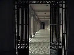 Galería techada vista a través de una puerta de rejas. A la izquierda se ven unas columnas. En el fondo se ve una puerta de madera.