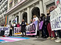 Concentración de mujeres trans en Madrid con una bandera del colectivo en el suelo (con blanco y tonos rosa y azul pastel), carteles reclamando igualdad y Mar Cambrollé, portavoz de laAsociación de Transexuales de Andalucía- Sylvia Rivera, en el centro pronunciando un discurso, a 9 de enero de 2024.