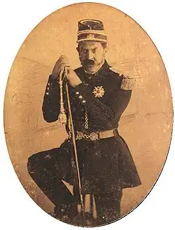 Three quarters length tintype portrait showing a mustachioed man in military dress uniform and cap with one foot resting on a rock and holding a sheathed sword.