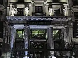 Fotografía de la entrada del Edificio Tornquist tomada de noche con iluminación ambiental que realza los rasgos de los ornamentos del frente del edificio. Se pueden apreciar cuatro pilastras, un cartel con el nombre del edificio y parcialmente su interior iluminado.