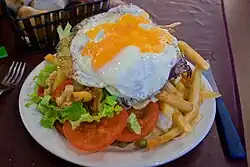 En la foto, se observa un chivito al plato con papas fritas, huevo frito, lechuga, tomate, pickles y arvejas. El chivito ocupa la totalidad del plato.