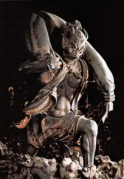 Fūjin. Three-quarter view of a statue. His left leg is bend as if climbing stairs and he is carrying a long bag-shaped object which goes from one shoulder to the other around the back of his head. Black and white photograph.