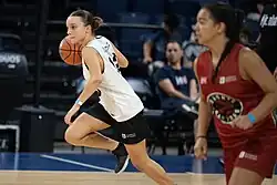 En un partido de básquetbol, dos mujeres jóvenes en primer plano, de distintos equipos, una de ellas lleva la pelota