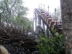 Una montaña rusa sube por encima de la vegetación. A la izquierda, la cabeza gris de un águila con una criatura ficticia.