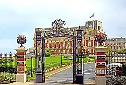 Photo couleur d'une grille ouvragée ouvrant sur un espace qui mène à un grand bâtiment rouge orné de jaune