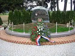 Un momument commémoratif, avec des inscriptions en serbo-croate et en allemand à la mémoire de la "tragédie de Bleiburg" ; devant le momument, une gerbe de fleurs aux couleurs du drapeau croate.