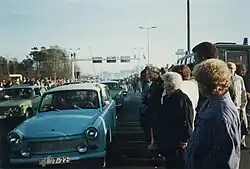 Coches Trabant de Alemania Oriental conducción entre densas multitudes de personas. Pórticos metálicos sobre la carretera y una torre de vigilancia son visibles en el fondo.