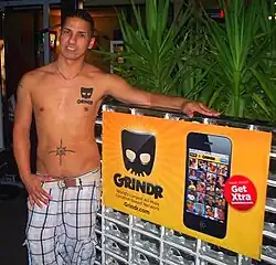Un hombre musculoso con el torso desnudo posa al lado de un cartel publicitario de la aplicación Grindr.