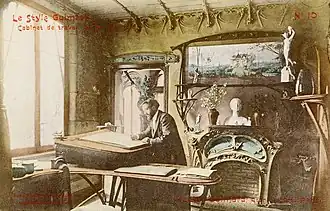 Photo sépia colorisée par endroits montrant un homme barbichu à un bureau dans un décor chargé