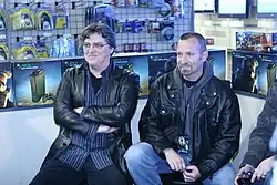 Two men wearing black leather jackets are sitting in chairs, surrounded by Halo-themed Xbox 360s and accessories. One person with the same attire is cutoff from the picture.
