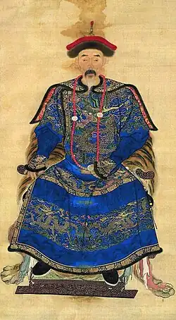 Un retrato en blanco y negro de un hombre sentado, con rostro demacrado, vistiendo una túnica cubierta con intrincados patrones de nubes y dragones.