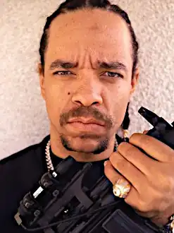 A man in his 40s, wearing a black top and several items of gold jewellery. He is holding what appears to be a large gun.