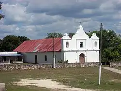 Vista sobre un campo abierto con parches de hierba gastada extendiéndose hacia una iglesia baja con una sencilla fachada blanca. La fachada tiene tres agujas cortas, la del medio más alta que las demás. La aguja central soporta una cruz. Las agujas más cortas cuentan con una abertura en forma de arco, una campana es visible en el arco de izquierda. La fachada cuenta con una puerta arqueada de madera; una ventana cuadrada está por encima de ella. La estructura principal de la iglesia tiene aproximadamente la mitad de la altura de la fachada y se extiende hacia atrás por el lado izquierda. La pared está lisa con excepción de una puerta más pequeña y una pequeña ventana. El techo es de metal corrugado oxidado. A la izquierda de la iglesia se encuentra un edificio bajo con una terraza sombreada. Dos postes soportan una línea eléctrica que pasa a través del campo a la iglesia. La base de cada poste está pintado de blanco. Una línea de árboles corre detrás de la iglesia.
