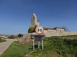 Imagen desde el inicio del pueblo donde puede verse el cartel con el nombre del municipio (Olmillos de Muñó) y, al fondo, el campanario de la iglesia La Asunción de Nuestra Señora)