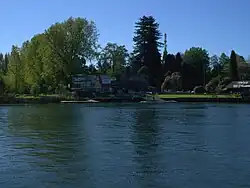 se ve el rio al frente, un muelle grande, pasto detras, una edificacion, y un par de carteles publicitarios junto a logo del club