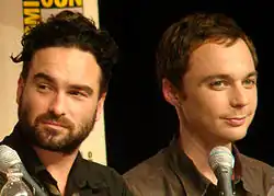 Two caucasian men, one with a beard, sit behind microphones, with a vague symbol in the background.