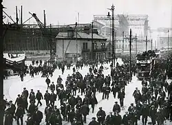 Trabajadores de Harland and Wolff a la salida del astillero, hacia abril de 1911. A la izquierda, se observa el SS Nomadic, transbordador del Olympic y del Titanic, durante su acondicionamiento. Al fondo, se observa el pórtico Arrol, con la proa del Titanic perfectamente visible.