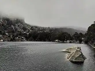 El agua contiene decenas de ondas provocadas por el viento. Al fondo de la foto se ve la cima de la montaña tapada por la niebla, que desciende hasta casi tocar el agua. La ladera de la montaña está manchada por trozos de nieve.