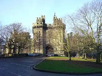 View down a short tree-lined drive leading to the main entrance of a castle, which is flanked by two tall towers.