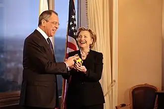 Hillary Clinton standing with Russian Foreign Minister Serguéi Lavrov. Both of them are holding a "reset button". They are in a room with a window to the left and an American flag behind them