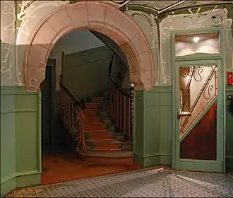 Photo en couleur d'un espace avec une ouverture en arc de cercle donnant sur un escalier et une porte vitrée surmontée d'arabesques peintes