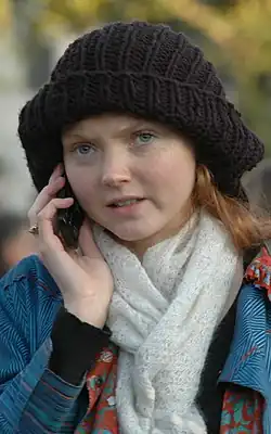 the head and shoulders of Cole outside on a mobile phone wearing a black beret, blue coat and white scarf